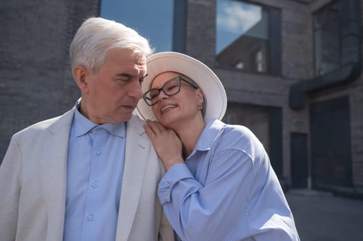 A woman in a hat and glasses hugs an elderly man in a white jacket from behind. Romantic relationships of mature people