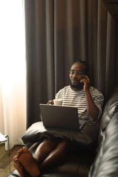 African American man freelancer talking on mobile phone and using laptop in living room.