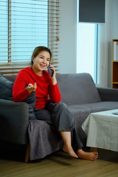 Cheerful young woman having phone conversation sitting on sofa at home.