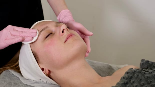 A professional cosmetologist cleans the client's face with a tonic with cotton pads before procedures. Vertical