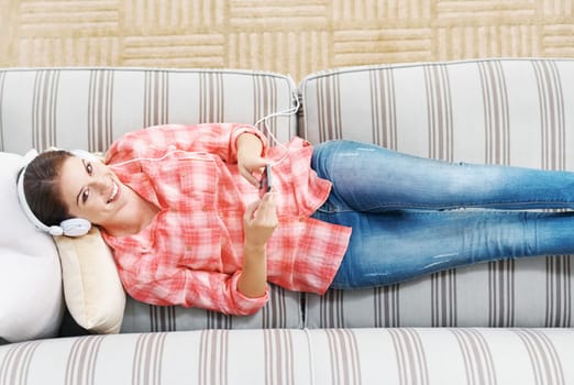 Happy woman, portrait and headphones with phone above in relax on sofa for music, podcast or audio streaming at home. Top view of young female person with smile on mobile smartphone for entertainment.