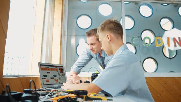 Smart student present coding program and car model to skilled teacher. Teenager using computer to programing system for robotic machines at table with electronic equipment scatter around. Edification.