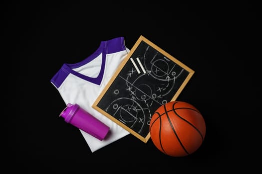 A basketball, along with a strategic play diagrammed on a chalkboard, rests near a folded jersey and a sports bottle. The equipment is arranged on a dark surface, suggesting preparation for a game or a coaching session in progress.