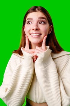 A cheerful young woman is standing against a vibrant green backdrop. Dressed in a cozy white sweater and gray pleated skirt, she playfully pulls at her collar with both hands. Her fashion-forward outfit is complemented by a pair of comfortable white socks, and her bright smile radiates a sense of lively confidence.