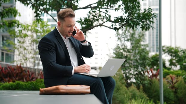 Caucasian businessman sitting at eco park while talking by using phone and working on laptop. Professional executive manager explain marketing idea from computer while calling to investor. Urbane