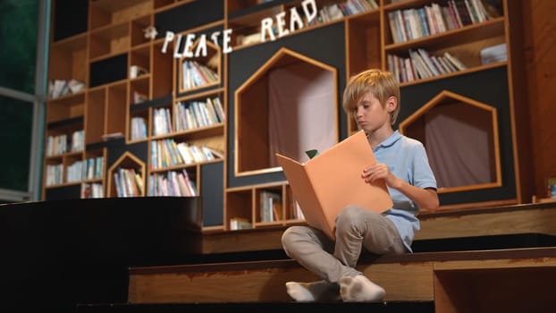 Attractive caucasian clever boy reading a book at library with stack of books placed. Smart child focus on studying, learning from novel or textbook while turning a page. Self-education. Erudition.