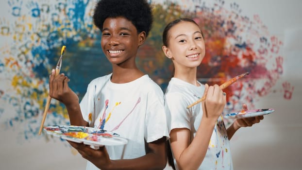 Highschool student with mixed races standing back to back and looking at camera at art lesson. Diverse children hold the paintbrush while pose with confident at colorful stained wall. Edification.