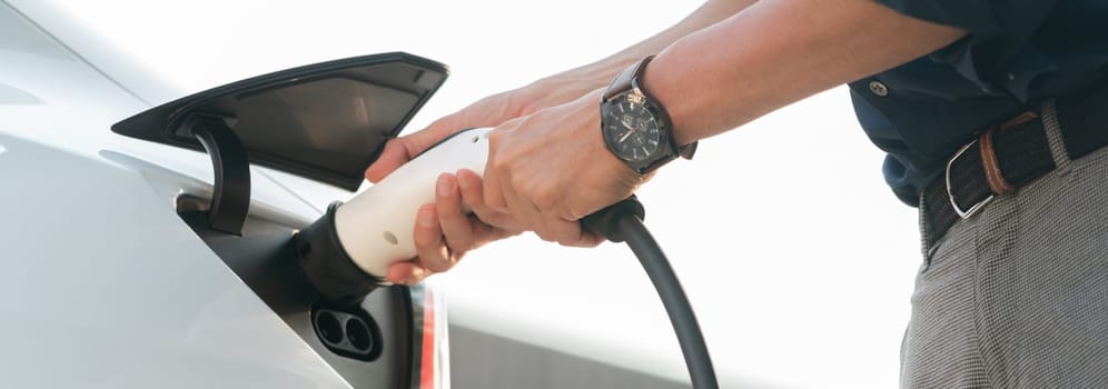 Young man put EV charger to recharge electric car's battery from charging station in city commercial parking lot. Rechargeable EV car for sustainable environmental friendly urban travel. Expedient