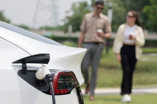 Young couple with coffee cup, recharge electric car's battery from EV charging station in green city park. Sustainable and eco friendly EV car with urban and shopping lifestyle. Expedient