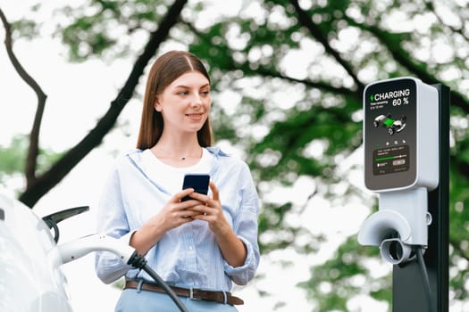 Young woman using smartphone online banking application to pay for electric car battery charging from EV charging station during vacation holiday road trip at national park or summer forest. Exalt