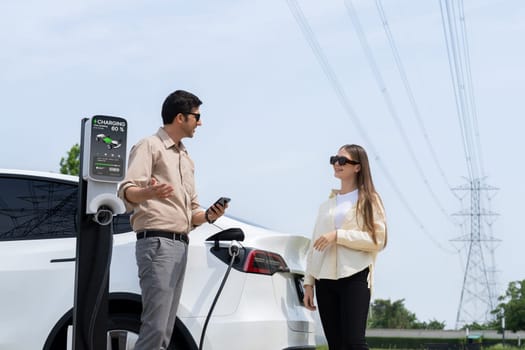 Couple pay for electricity with smartphone while recharge EV car battery at charging station connected to power grid tower electrical as electrical industry for eco friendly car utilization.Expedient