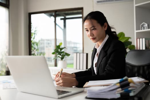 Focused Asian female accountant doing paperwork in office with plan documents on desk.