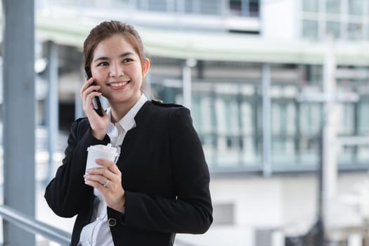 Asian businesswoman drinks coffee and discusses work on the phone in the hallway outside the office..