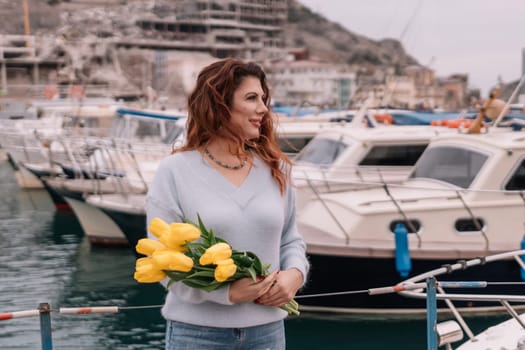 Woman holds yellow tulips in harbor with boats docked in the background., overcast day, yellow sweater, mountains.