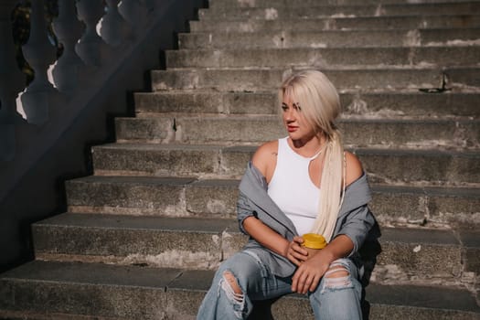 A blonde woman sits on a set of stairs, holding a cup. She is wearing a gray jacket and blue jeans
