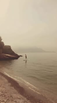 A person is balancing on a paddle board in the calm waters of a serene lake with a picturesque landscape of trees, sky, and clouds in the background