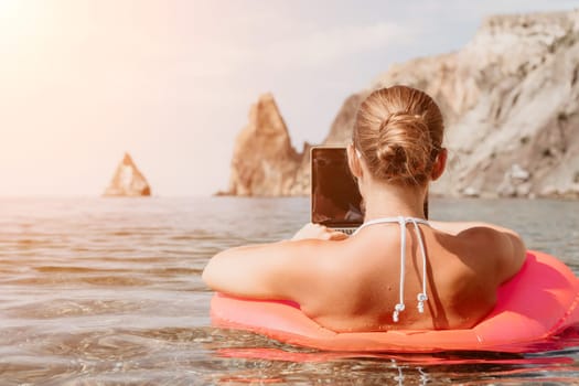 Woman freelancer works on laptop swimming in sea on pink inflatable ring. Pretty lady typing on computer while floating in the sea on inflatable donut at sunset. Freelance, remote work on vacation