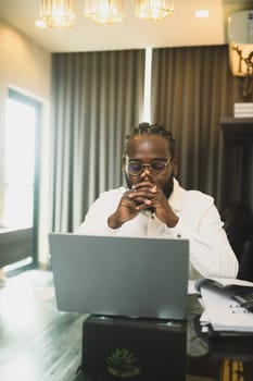 Serious African American man entrepreneur sitting in front of laptop with hands clasped.