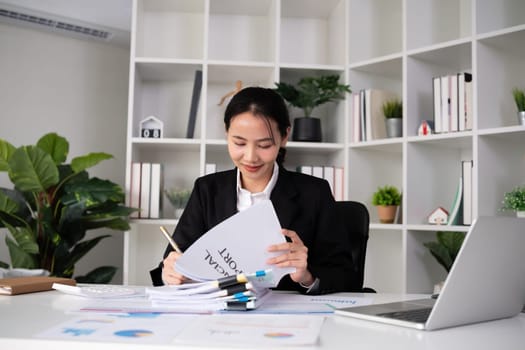 Focused Asian female accountant doing paperwork in office with plan documents on desk.