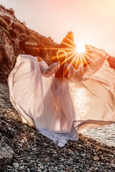 Mysterious woman silhouette long hair walks on the beach ocean water, sea nymph wind listens to the wave. Throws up a long white dress, a divine sunset. Artistic photo from the back without a face.