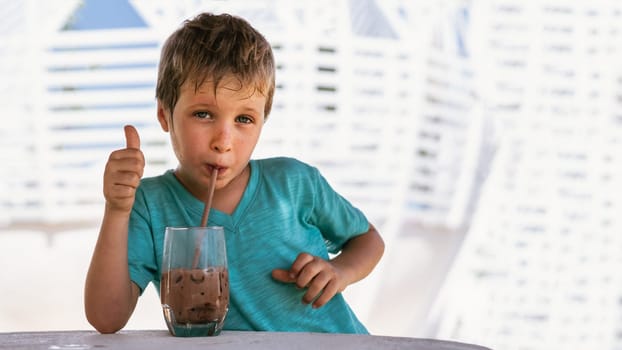 Happy childhood. Boy drinking chocolate tea with ice in hot summer day time. Cute lover of sweets and tasty things. Shows thumbs up.