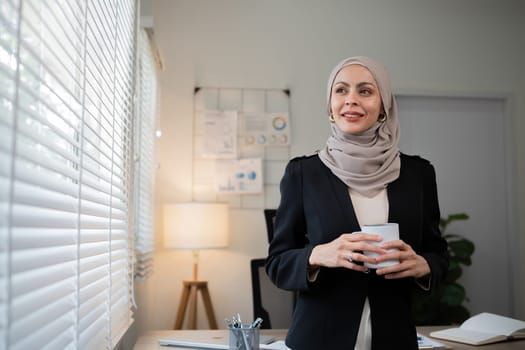 Confident Muslim businesswoman standing with coffee mug in front of desk in office during morning time.