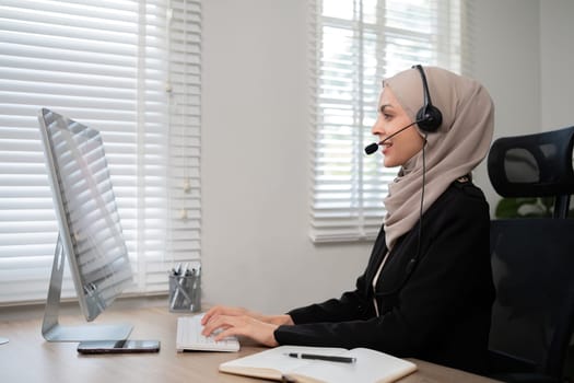 Call center worker, young Muslim woman wearing hijab, talking to customer on call phone on computer in customer service office.