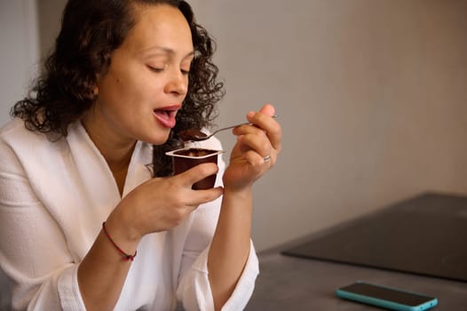 Close-up portrait of a multi ethnic beautiful young woman at home eating yoghourt for breakfast, dressed in white bathrobe in the morning