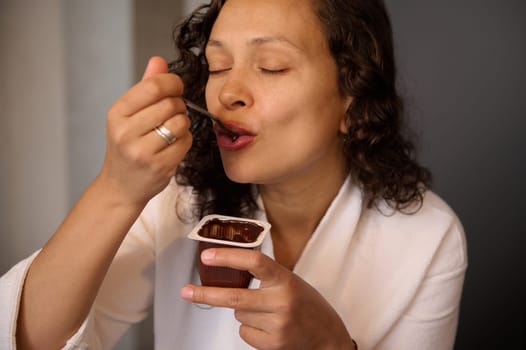 Authentic portrait of curly haired brunette woman enjoying the taste of chocolate vegan yogurt. Attractive young woman with closed eyes, dressed in white bathrobe taking healthy yoghurt for breakfast