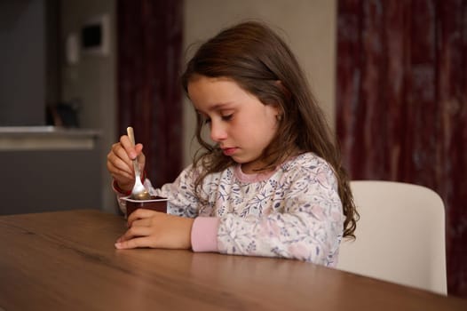 Caucasian adorable little child girl eating delicious chocolate vegan yogurt for breakfast. Healthy eating and diet concept. Food and drink consumerism.