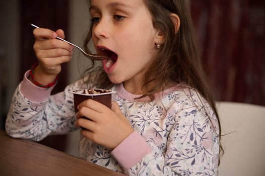 European little kid girl snacking with a tasty yoghurt at home. Authentic portrait of a lovely child 6 years old, enjoying the taste of chocolate vegan yogurt. Food and drink consumerism. Healthy diet