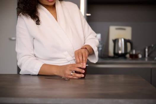 Close-up of a young curly hired multi ethnic woman in white bathrobe, standing at kitchen table at home, having a chocolate yoghourt for breakfast. Domestic life.