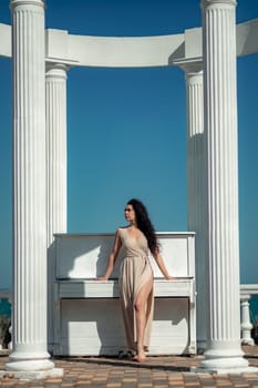 A woman in a long dress stands in front of a piano. The piano is white and has a few keys visible. The woman is posing for a photo, and the overall mood of the image is calm and serene