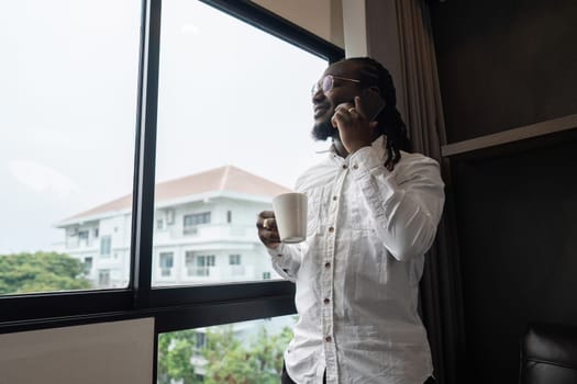 African American man using have call by phone while drink coffee by the window in the living room.