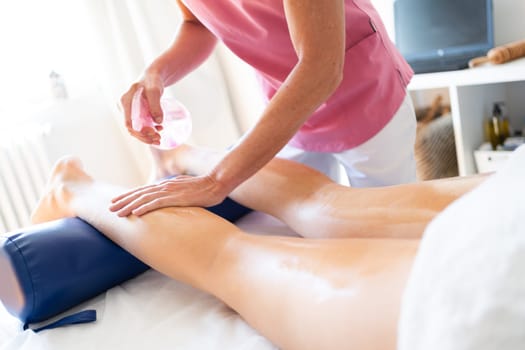 Crop anonymous massage therapist using oily spray cream while massaging leg of client lying down on bed during physiotherapy session in clinic