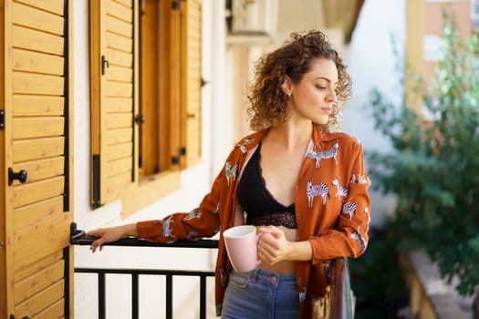 Young stylish female in casual clothes with curly hair standing on balcony and holding cup of coffee while looking down
