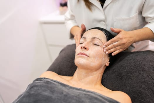 From above crop cosmetician massaging face of young female client with lotion during facial treatment in beauty salon on blurred background