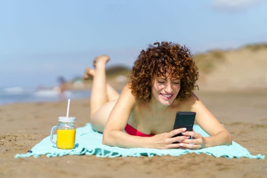 Young curly haired female in bikini lying on sandy beach and browsing mobile phone while having cup of orange juice