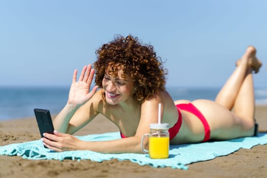 Happy curly female tourist lying on sandy coast near sea in summer and waving hand while chatting online via cellphone