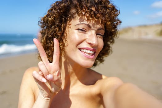 Cheerful young female with curly hair smiling and looking at camera while showing two fingers and taking selfie on sandy beach on sunny day