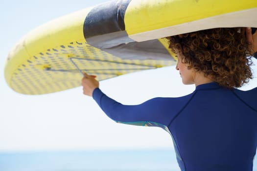 Back view of crop anonymous female with curly hair in casual clothes standing with raised SUP board against blue sea background on sunny day