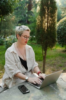 woman freelancer in glasses works at a computer at a white table in nature and spends her day productively.