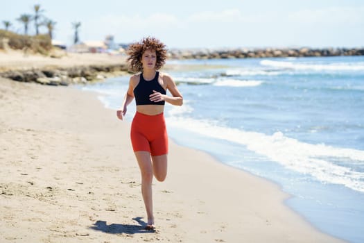 Full body of determined young female with curly hair in orange shorts and black top running along sandy seashore on sunny day