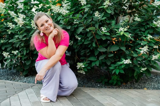 Portrait of beautiful smiling millennial woman wearing stylish pink t shirt looking at camera standing in green park. Positive lifestyle, natural beauty concept. Copy space.