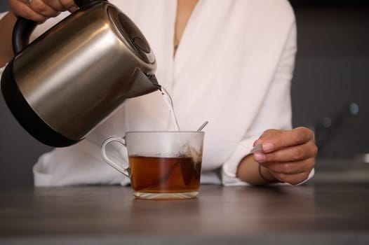 Close-up of a woman making tea for breakfast. Selective focus on female hands pouring boiling water from an electric stainless steel teapot into glass cup with tea bag. Food and drink consumerism