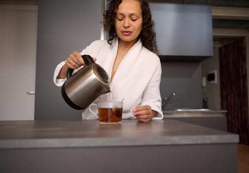 Young pretty woman making tea for breakfast. Selective focus on female hands pouring boiling water from an electric stainless steel teapot into glass cup with tea bag. Food and drink consumerism