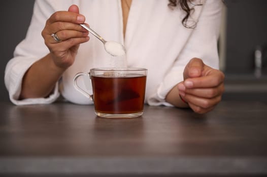 Close-up of woman's hands holding a teaspoon with sugar, sweetening the drink in a glass. Woman's hands holding a spoon with sugar. Adding sugar to a cup of hot drink beverage