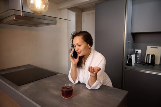 Multi ethnic attractive woman in white bathrobe talking on mobile phone while having light breakfast in the home kitchen. People. Morning routine, Lifestyle