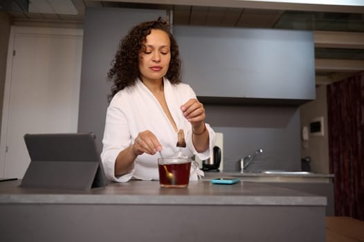 Confident pretty brunette woman in white bathrobe, sweetening his tea, standing at kitchen table with open digital tablet, in a minimalist home kitchen interior