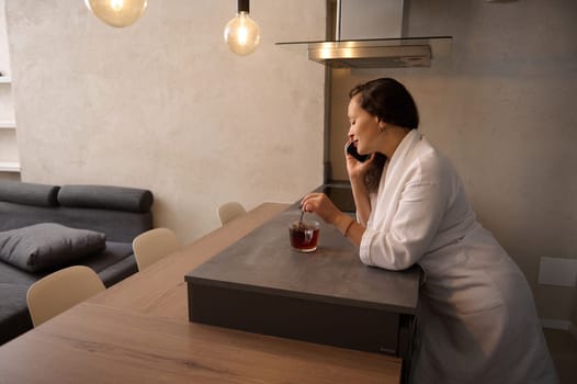 Relaxed brunette in white bathrobe, talking on smartphone, standing at kitchen table and mixing sugar in cup of hot tea. Attractive woman having a conversation on telephone in minimalist home interior